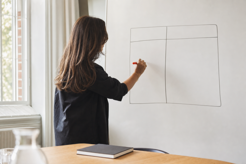 A marketing strategist, seen from the side, is drawing a two-column comparison on a whiteboard with coral-colored marker in a brightly lit conference room